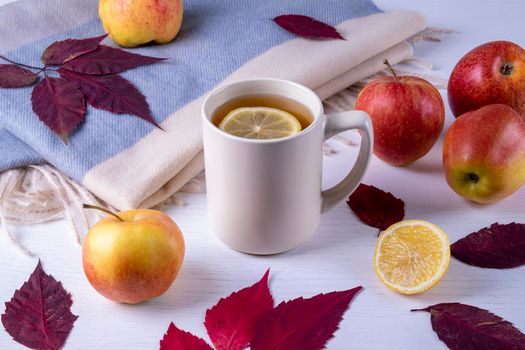 Autumn composition of ceramic mug with hot tea with lemon, soft warm scarf in pastel colors, dry autumn leaves and ripe apples. Selective focus.