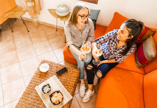 Couple of young women friends drinking at home sitting at orange sofa celebrating with champagne or white wine. Girls using alcohol to having fun taking aperitif in living room instead of study lesson