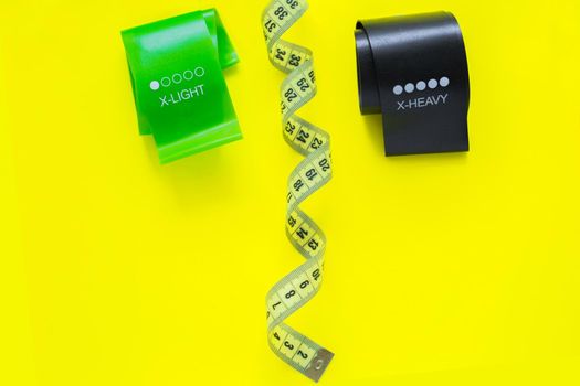 Two fitness tapes of black and green color are located at the top of the photo on a yellow background, they are separated by a yellow measuring tape.