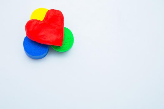 A heart made of red dough for modeling lies on top of a yellow, green and blue mug on a white background. High quality photo