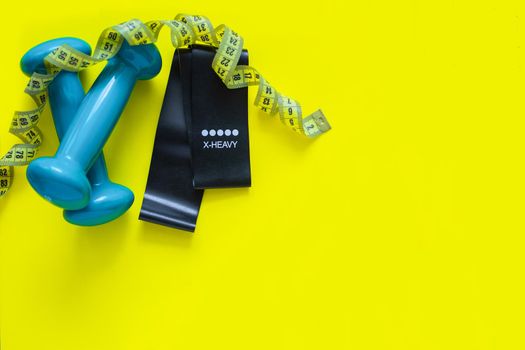 A black fitness elastic band and two blue dumbbells lie on a yellow background, a measuring tape lies above them in rings, there is a place for text.