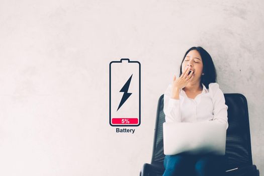 Young asian woman yawn at her work place with laptop computer on cement background, girl working with yawn and burnout with symbol battery low, stress and tired, copy space, business concepts.
