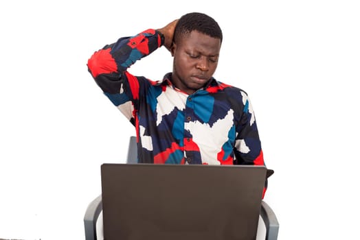 young african sitting on chair in multicolored shirt looking at laptop.