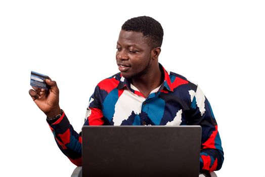 young african sitting in multicolored shirt looking at credit card smiling with laptop on thighs.