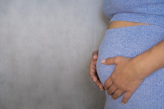 A pregnant woman is standing in a blue suit and holding her tummy with her hands on a gray background. High quality photo