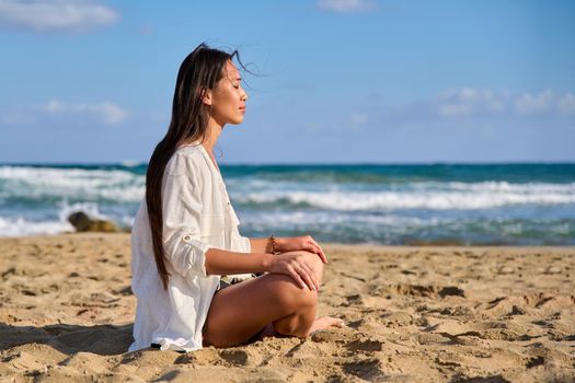 Young beautiful woman in lotus position meditating on the beach. Relaxing resting Asian female with closed eyes sitting on sand, scenic seascape background. Lifestyle, health, beauty, nature, people