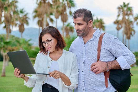 Middle-aged man and woman looking in laptop screen outdoors. Couple of business colleagues talking discussing smiling, tropical park nature background. Business, people, technology, mature age concept