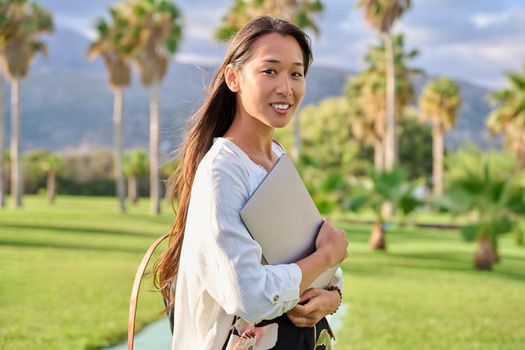 Outdoor portrait of young woman with laptop and backpack. Asian female adult student, creative freelancer, in tropical park, mountains background. E-learning, online technologies, education, business