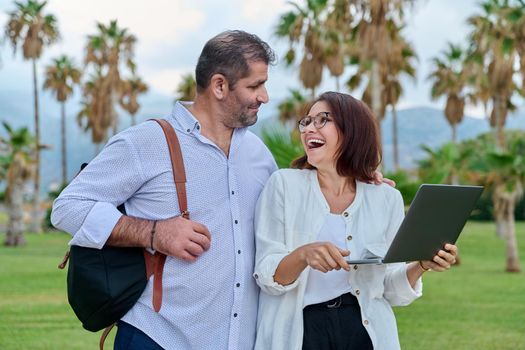 Middle-aged man and woman looking in laptop screen outdoors. Couple of business colleagues talking discussing smiling, tropical park nature background. Business, people, technology, mature age concept