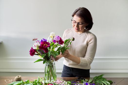 Mature woman at home with spring flowers. Female making bouquet, placing flowers in jug of water. Spring summer season, floristry, home decoration, natural beauty concept