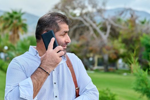 Mature business man talking on the phone outdoors. Serious confident male with backpack, in tropical park. Business trip, freelance, communication, technology, middle aged people concept, copy space