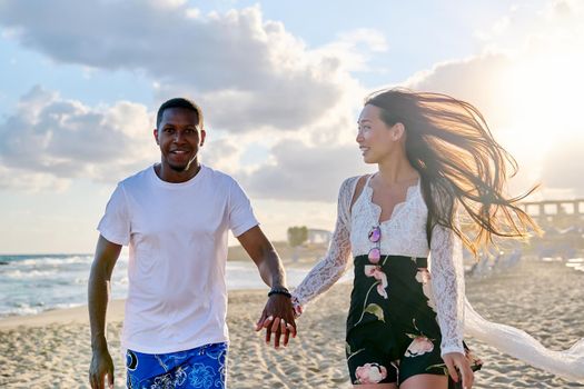 Beautiful young couple running along beach together. Happy loving african man and asian woman holding hands. People, love, happiness, relationship, multiethnic family, vacation, tourism