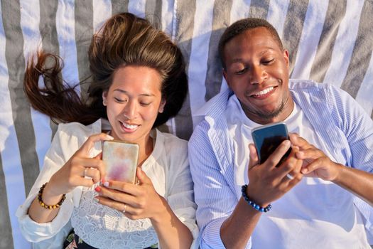 Top view smiling young couple looking at smartphone screens. Interracial couple of asian woman and african man outdoor vacationing, lying with phones in hand. Lifestyle, leisure, technology, people