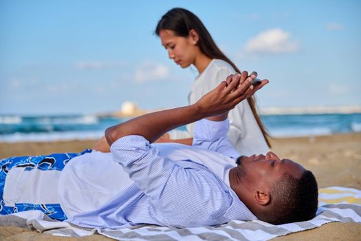 Young beautiful multiethnic couple having a rest on the beach, African American man with a smartphone in his hands close-up. Lifestyle, technology, leisure, people concept