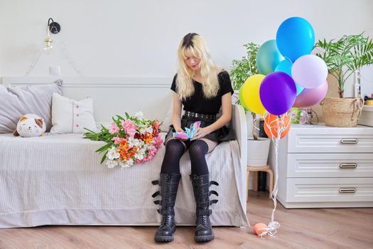 Birthday of teenage female 16, 17 years old. Hipster teenager at home with surprise gift in box, bouquet of flowers, colored balloons, sitting at home on bed. Teens, age, holiday concept