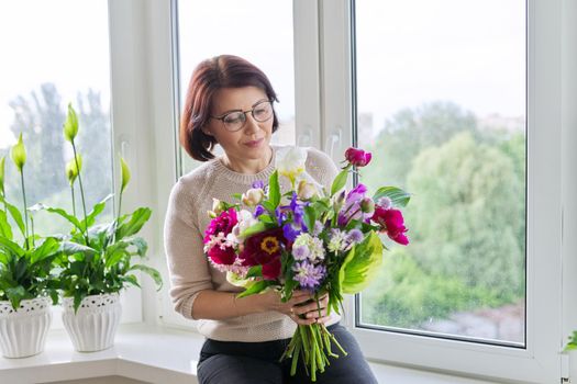 Portrait of beautiful mature woman with bouquet at home. Smiling female with flowers and buds of peony iris lupine. Spring summer season, natural beauty, middle aged people, gift, holiday concept