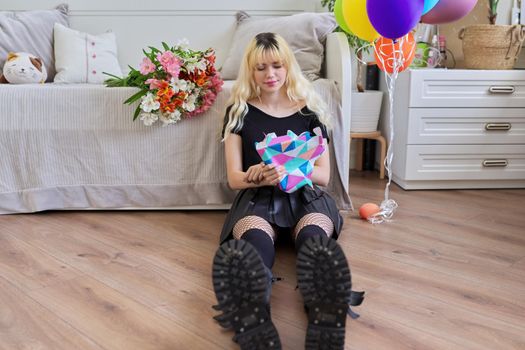 Birthday of teenage female 16, 17 years old. Hipster teenager at home with surprise gift in box, bouquet of flowers, colored balloons, sitting at home on bed. Teens, age, holiday concept