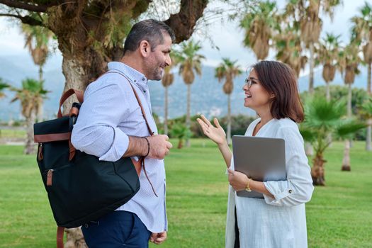 Mature man and woman business colleagues talking outdoors. Female and male together discussing, smiling, debating, standing in tropical park. Business, people, team, teamwork, middle age concept