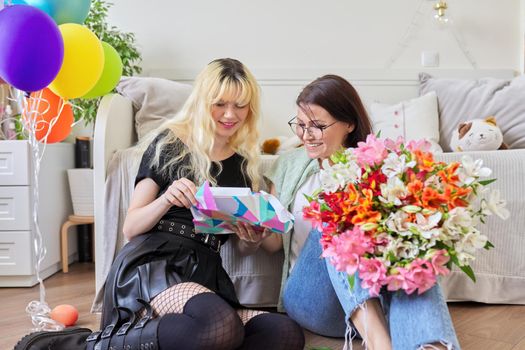 Mother and teen girl, happy birthday. Female mom congratulating daughter with bouquet of flowers and surprise gift box, room interior background. Holiday, celebration, parent teenager relationship