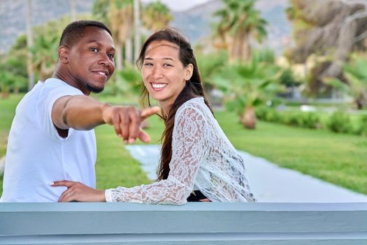 Beautiful young multi-ethnic couple sitting together on bench in tropical park. Loving asian woman and african man embracing. Male showing index finger. Relationship, happiness, multicultural family