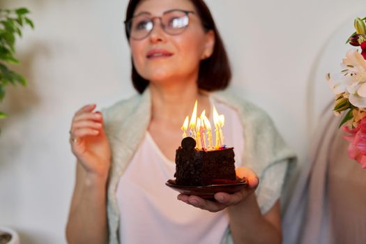 Mature woman with small birthday cake with burning candles. Happy 40s woman making wish before blowing out the candles. Age, birthday, anniversary, joy holiday happiness concept