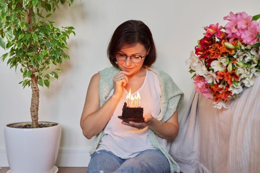 Middle-aged women with small birthday cake with candles. Happy female at home on the floor with bouquet of flowers. Age, date, aging, celebration, lifestyle, anniversary concept