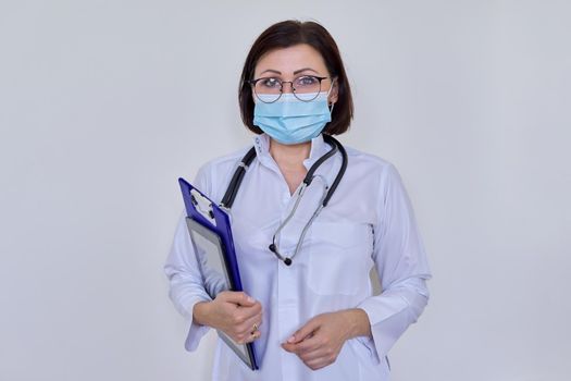 Portrait of female doctor in white uniform in protective medical face mask with stethoscope, with clipboard and digital tablet in her hands, woman looking at camera, on light background