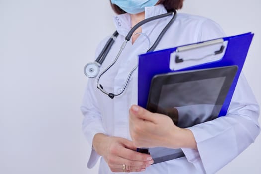 Female doctor in white uniform in protective medical face mask with stethoscope with clipboard and digital tablet in her hands, on light background, copy space