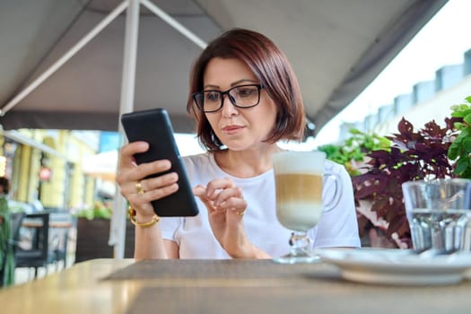 Serious middle-aged woman sitting in an outdoor cafe with a cup of coffee and looking at the smartphone screen. Coffee break, people 40s age, urban style, leisure, lifestyle, technology concept