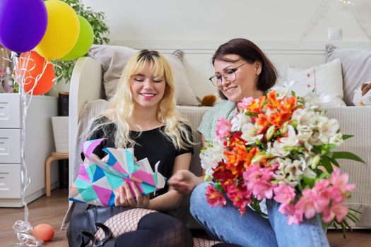 Mother and teen girl, happy birthday. Female mom congratulating daughter with bouquet of flowers and surprise gift box, room interior background. Holiday, celebration, parent teenager relationship