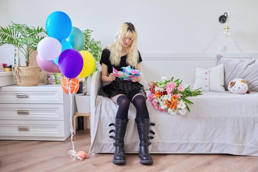 Birthday of teenage female 16, 17 years old. Hipster teenager at home with surprise gift in box, bouquet of flowers, colored balloons, sitting at home on bed. Teens, age, holiday concept