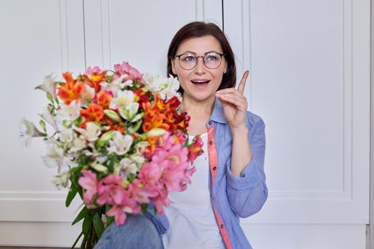 Portrait of beautiful mature woman with bouquet of flowers, at home against the background of light wall. Age, middle-aged people, holiday, beauty, date, anniversary, greetings concept