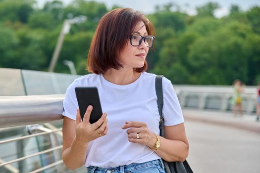 Portrait of middle-aged woman in glasses jeans white T-shirt walking with smartphone in hand, summer day in city. Lifestyle, urban style, middle-aged people concept