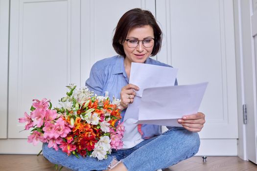 Middle-aged smiling female with a bouquet of flowers reading paper. Holiday, date, anniversary, good business news, positive letter, congratulations concept