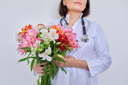 Close-up of bouquet of flowers in hands of female doctor. Medic, pharmacist in white uniform with stethoscope on light background. International, National Doctor's Day, Nurse's Day, Health Day