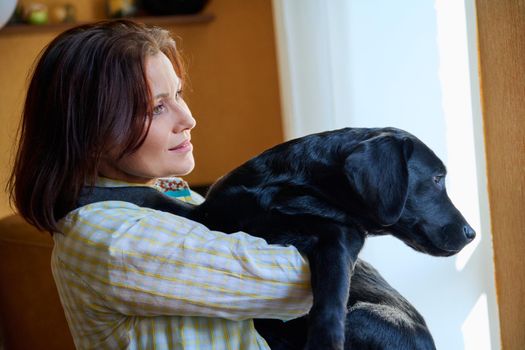 Portrait of middle aged woman and black labrador puppy dog. The owner holding young pet in her arms, in home interior. Lifestyle, love, pets, 40s people concept