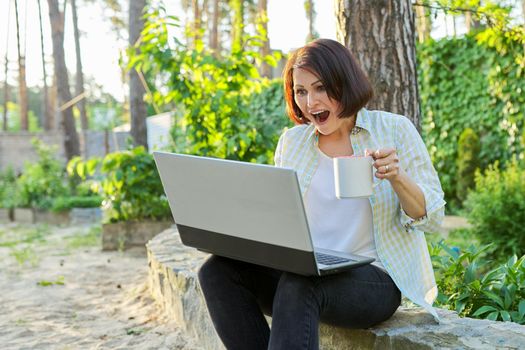 Beautiful emotional middle aged woman relaxing in garden with cup of tea and laptop. Female using video call for virtual meeting, watching movies, reading, having fun