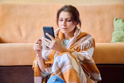 Autumn winter portrait of middle aged woman under warm blanket with hot cup of tea and smartphone in hands. Female resting at home, sitting on the floor near the sofa