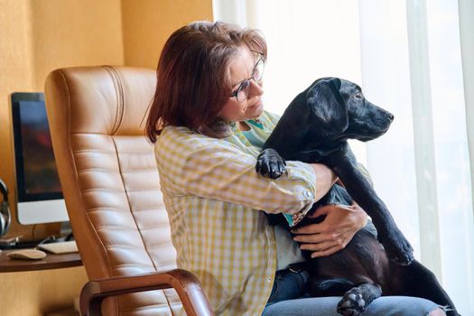 Portrait of middle aged woman and black labrador puppy dog. The owner holding young pet in her arms, in home interior. Lifestyle, love, pets, 40s people concept