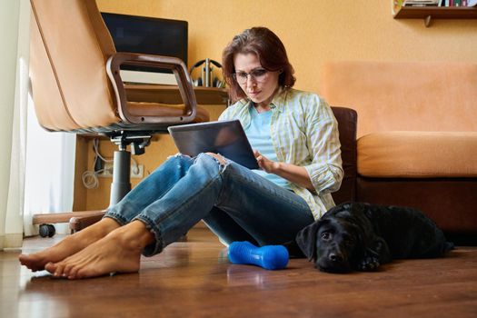 Middle-aged serious woman sitting at home on the floor with digital tablet, female working at home, dog lying resting next to owner