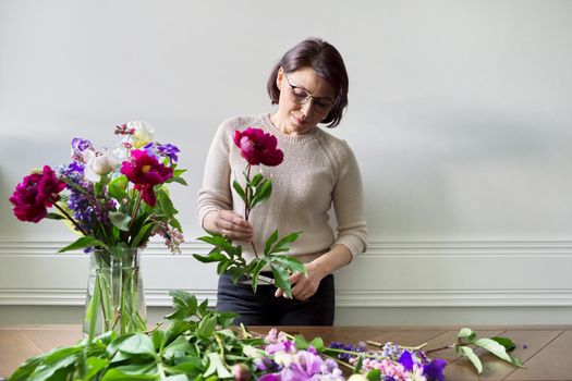 Mature woman at home with spring flowers. Female making bouquet, placing flowers in jug of water. Spring summer season, floristry, home decoration, natural beauty concept