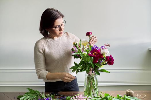 Mature woman at home with spring flowers. Female making bouquet, placing flowers in jug of water. Spring summer season, floristry, home decoration, natural beauty concept