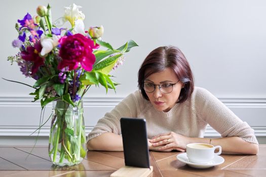 Serious mature woman looking into smartphone screen. Middle-aged female at home at table with smartphone, listening and looking attentively and intently