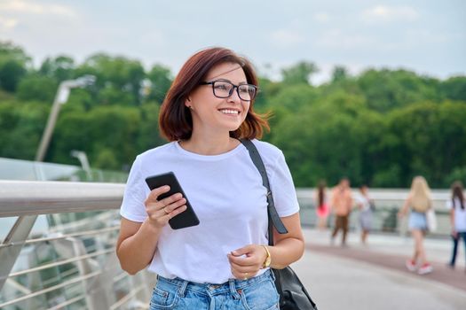 Portrait of middle-aged woman in glasses jeans white T-shirt walking with smartphone in hand, summer day in city. Lifestyle, urban style, middle-aged people concept