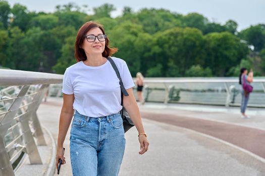 Portrait of middle-aged woman in glasses jeans white T-shirt walking with smartphone in hand, summer day in city. Lifestyle, urban style, middle-aged people concept