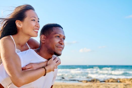 Young beautiful having fun couple on sea background. Multiracial couple, asian woman on back of african american man, vacation together, relationship dating happiness, happy lovers moments, copy space