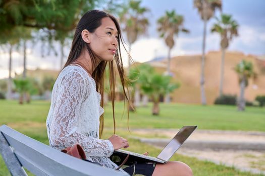 Young beautiful woman outdoors with laptop, tropical park background. Asian female sitting on bench, working, studying remotely. Freelance e-learning, online technologies, education, business concept