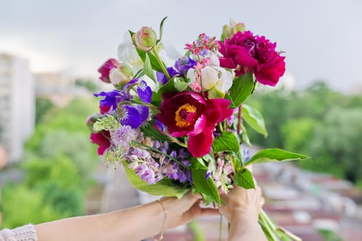 Close-up of bright bouquet of flowers in female hand. Fresh flowers and buds of peonies iris lupine in flower arrangement. Season spring summer, natural beauty, background open window blue sky