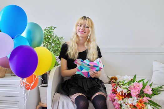 Birthday of teenage female 16, 17 years old. Hipster teenager at home with surprise gift in box, bouquet of flowers, colored balloons, sitting at home on bed. Teens, age, holiday concept