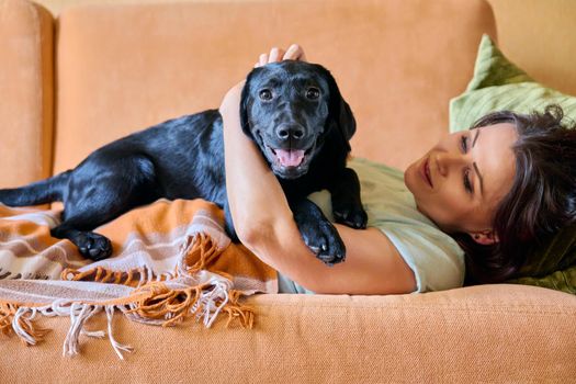 Relaxed middle aged woman lying on sofa under blanket and petting black labrador puppy dog. Lifestyle, love, care, pets, 40s people concept